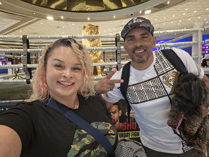 A man and woman pose for a selfie inside a boxing arena, with a golden lion statue and 'PREMIER BOXING CHAMPIONS' branded ropes visible in the background.