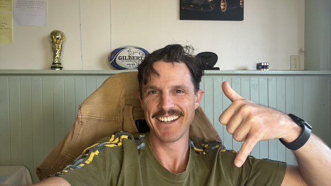 The image features a man sitting at his desk with a green shirt on, smiling and making a thumbs up gesture. The background of the photo shows a white wall with a trophy displayed behind him. There are also some papers visible on the wall. The caption reads "Join me from my desk in South Africa in just under an hour, peeps! Join me for a post-work beverage if you’re allowed to enter this draw to win $SUPRA LIVE ON THE SHOW."