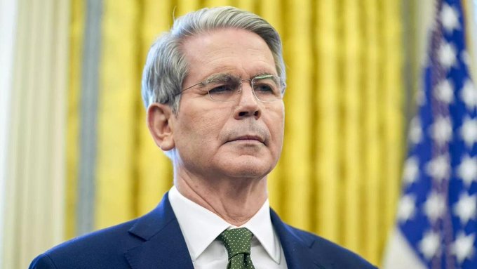 A portrait of a gray-haired man wearing glasses, a navy suit, and green tie, standing in front of yellow curtains and an American flag.