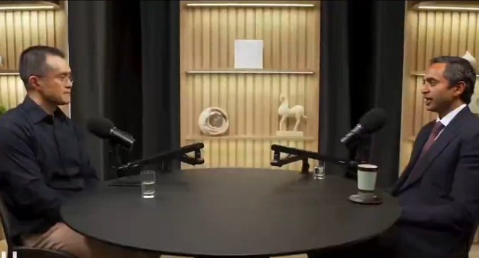 A podcast-style interview setting shows two men in suits seated across from each other at a round table with microphones, engaged in a formal discussion against a backdrop featuring a horse statue and decorative shelving. The image aligns with the post's context of CZ, the founder of Binance, sharing his early Bitcoin investment story during what appears to be a recorded conversation.