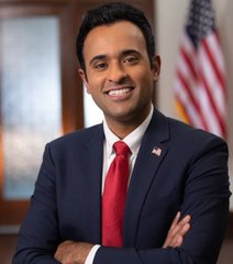 A professional portrait of a smiling man in a dark suit and red tie, standing with crossed arms in front of an American flag.