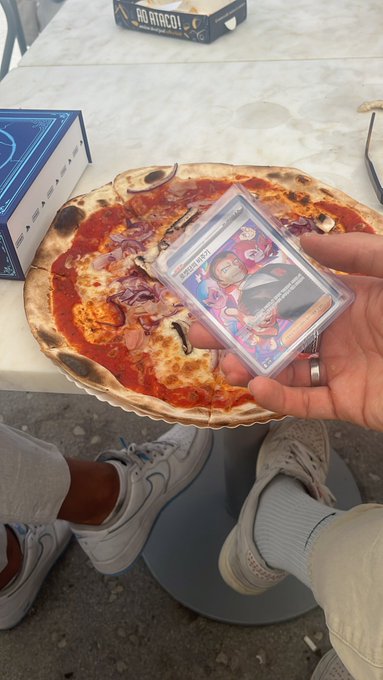 The image features a pizza with various toppings, including pepperoni and sausage, sitting on top of a table. A person's hand holding a Pokémon card can be seen next to the pizza. The post includes text that reads "got a pack from @StarPlatinum_ pulled a banger." This suggests that someone has been playing Pokémon or is discussing it with others who have played the game.