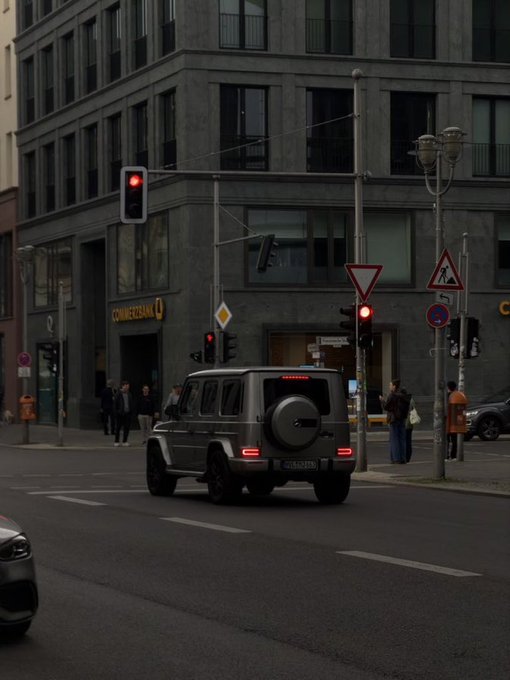 A street scene showing a gray Mercedes-Benz G-Class SUV at a red light in an urban area with a Commerzbank building visible.