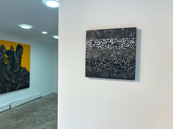 An art gallery interior featuring a black and white textured square painting on a white wall, with another colorful painting depicting elephants and figures visible in the adjacent room.