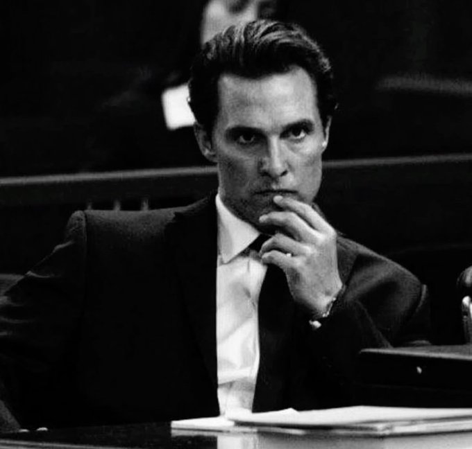 A black and white photo of a man in a suit and tie sitting at a desk with his hand near his chin, appearing thoughtful.