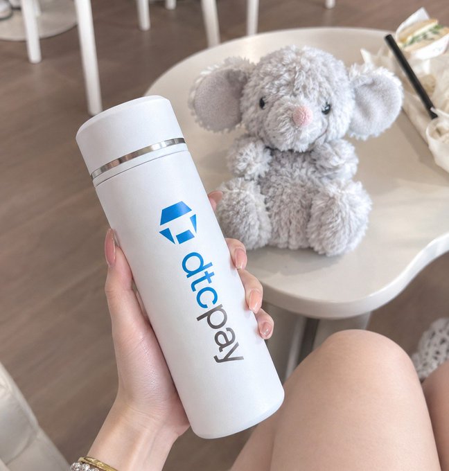 A hand holds a white thermos with the 'aicpay' logo next to a grey plush elephant toy sitting on a white table.