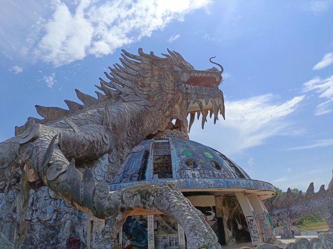 A massive dragon sculpture with detailed scales and menacing teeth towers over a dome-shaped building with mosaic decorations, photographed against a bright blue sky.
