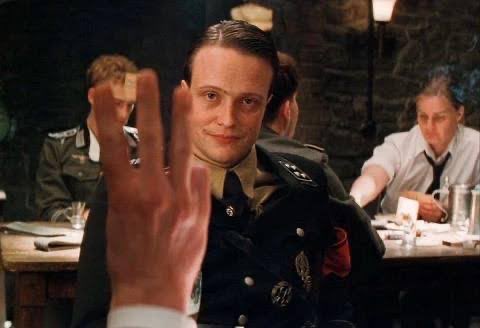 A man in a Nazi uniform gives the three-finger salute at a dinner table. The image relates to the post through a humorous reference to the "3" gesture and the infamous "Solana Nazi" meme, which has become a cultural shorthand in crypto circles for waving away speed claims people consider overblown or irrelevant.