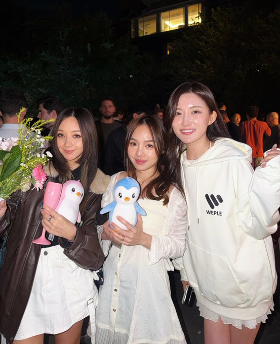 Three young women pose together at a nighttime outdoor event; the woman on the left holds flowers and a pink penguin figurine, the center woman holds a blue penguin figurine, and the woman on the right wears a cream hoodie with "WEPLE" text.