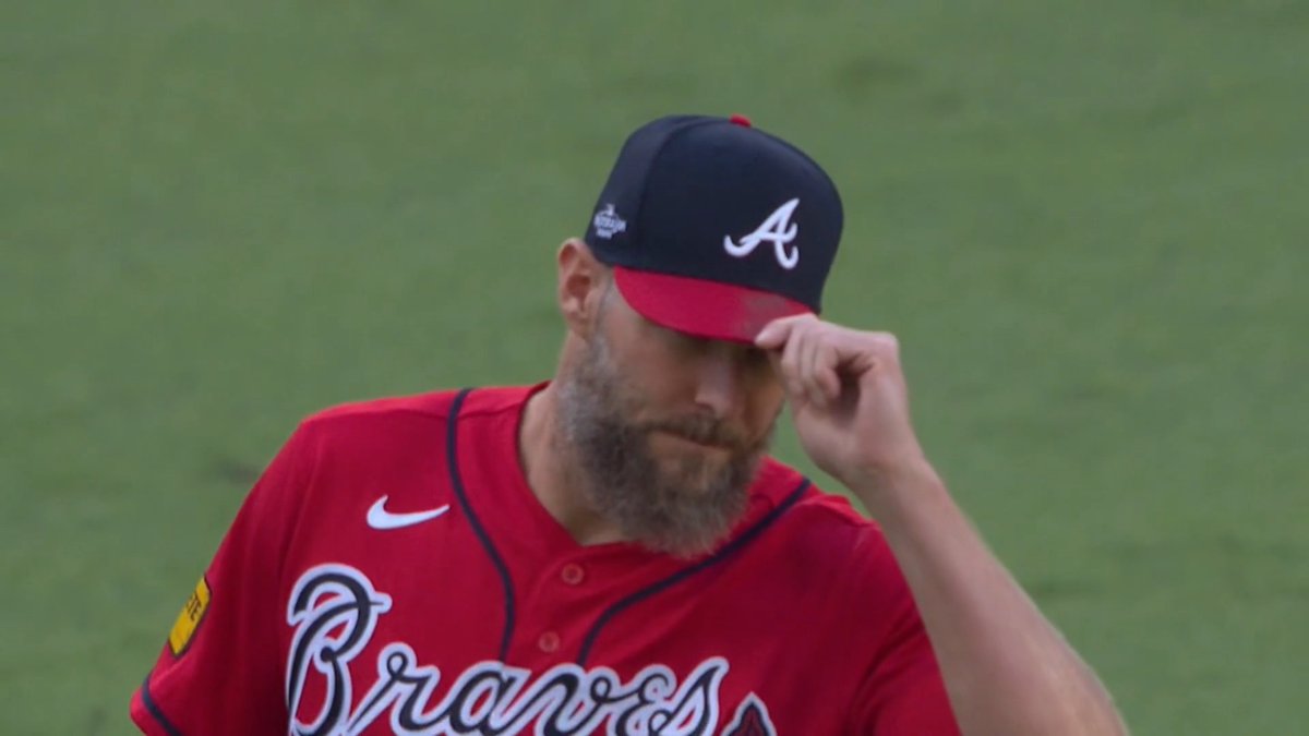 A bearded baseball player wearing a red Atlanta Braves jersey and a navy blue cap with a red brim, looking down while adjusting his cap on the field.