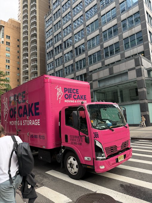 A street-level photograph of a bright pink moving truck for the company "Piece of Cake Moving & Storage" turning at a city intersection, with modern buildings in the background and pedestrians in the foreground.