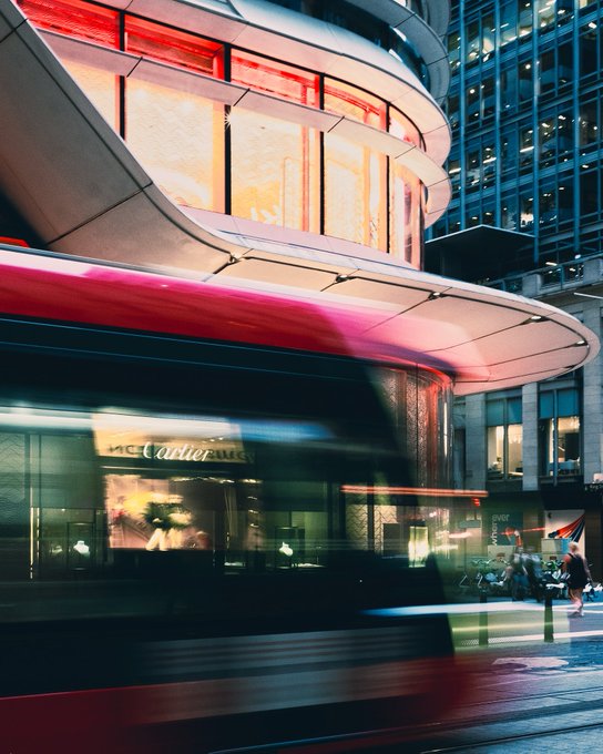 A motion-blurred nighttime photograph of a red double-decker bus passing a curved, illuminated building in an urban setting.