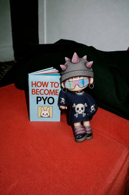 A toy figure with spiked pink horns and skull-patterned clothing stands beside a small book titled "HOW TO BECOME PYO" on a red couch, visually tying into the accompanying post about the PYO ecosystem report detailing soft staking milestones and $MTH token burns and distributions.