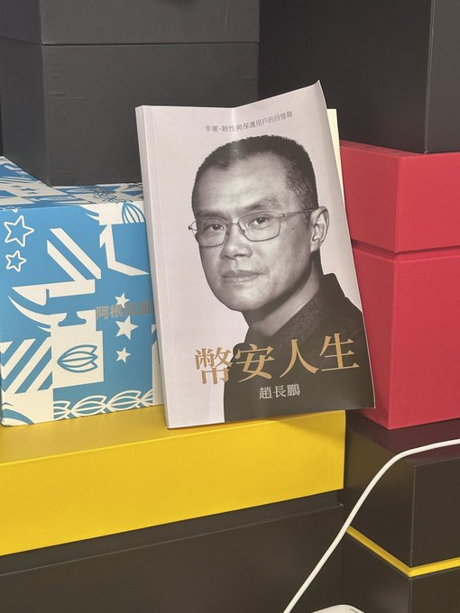 The image shows a book titled "幣安人生" (Binance Life) by 趙長鵬 (CZ) propped up against colorful stacked boxes, with the subtitle "幣圈·幣咖與保應用的思維" visible. This connects to the post's reference to @cz_binance, as it displays CZ's memoir, emphasizing the playful mention of paying "respects to Big Cousin-GM."