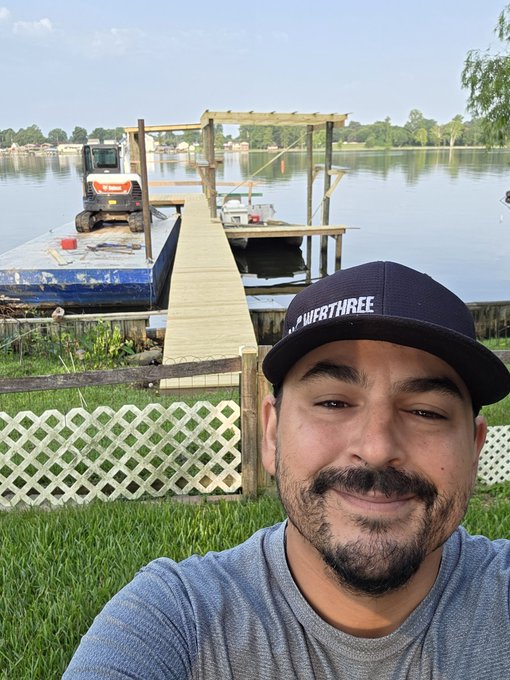 A man in a "Stay Classy NWA FORT THREE" snapback cap smiles in the foreground, with a newly constructed wooden pier extending from a lakeside dock behind him, where a small Bobcat skid-steer loader sits on a pontoon platform. The image captures the construction progress mentioned in the post, showing the Web3Auditing office's new pier nearly complete on the calm water, ready to host clients "the southern way."