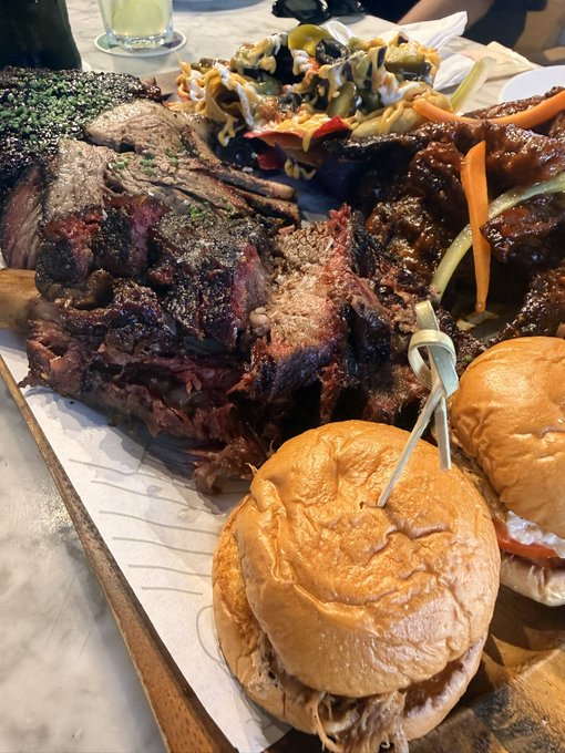 The image shows a large platter of food with two sandwiches placed next to each other on a wooden board. The sandwiches are made from pulled pork and include various toppings such as carrots and pickles, along with some sauce. In addition to the sandwiches, there is also a plate of meat covered in sauce nearby. A glass filled with a beverage can be seen at the top left corner of the image.