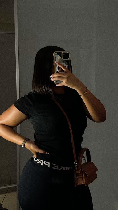 A mirror selfie of a woman wearing a black Nike t-shirt and black Nike Air leggings, carrying a brown crossbody bag while holding a phone in front of her face.