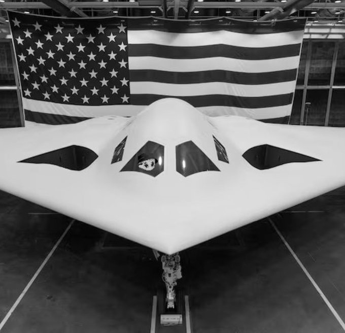 A black-and-white photo of a sleek, futuristic B-2 Spirit stealth bomber parked in a hangar with a large American flag hanging in the background.