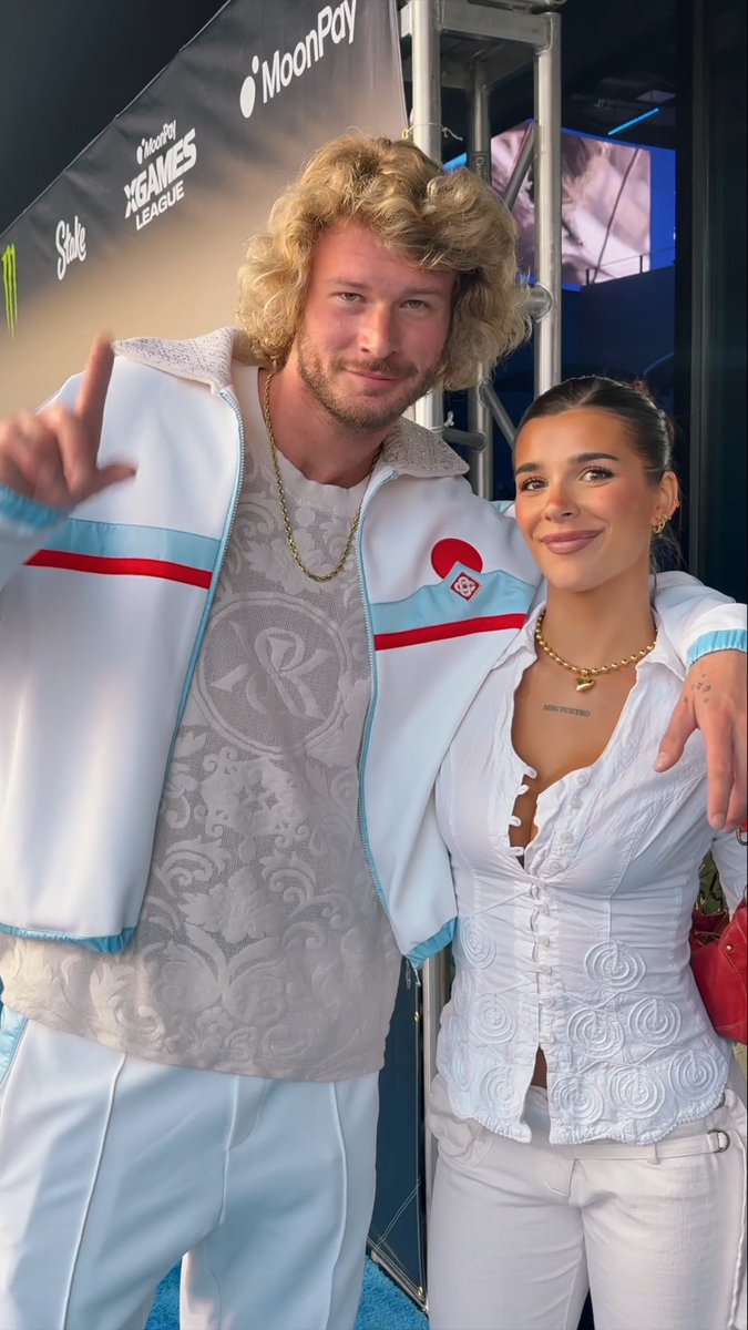 A curly-haired man and a brunette woman pose together on the blue carpet at the MoonPay X Games League Winter Draft, both dressed in coordinated white outfits featuring blue and red accents, with the event's sponsor logos visible in the background.