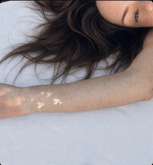 A woman with freckled skin and long brown hair lies on a white surface, showing several smears of beige liquid foundation swatched on her forearm as a beauty product test.

The image relates to the post by showing foundation swatches on the woman's arm, with the caption celebrating her "solana" (golden, sun-kissed) glow from Sardinia—likely referencing how the foundation matches her sun-tanned, freckled complexion.