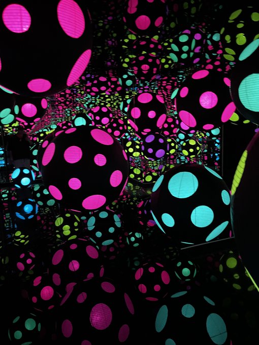 An art installation featuring glowing spherical orbs covered in pink, green, and cyan dots displayed in a dark mirrored room, characteristic of Yayoi Kusama's work.