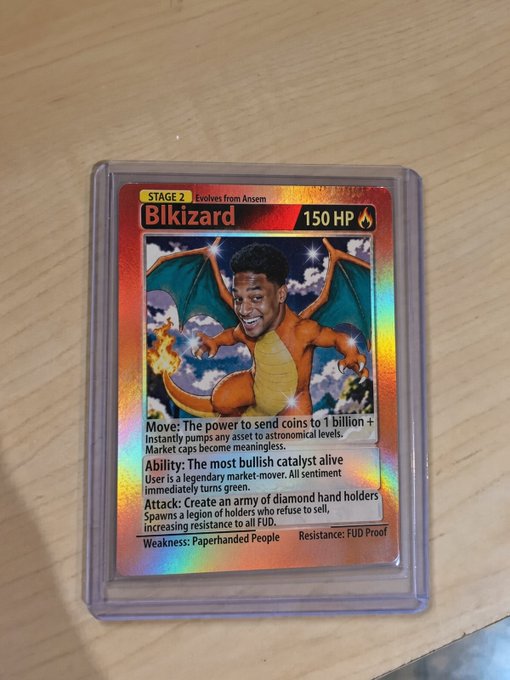 A photograph of the same custom 'Bikizard' Pokémon trading card (150 HP) lying on a light wooden surface, showing its detailed crypto-parody text including 'Move: The power to send coins to 1 billion' and 'Resistance: FUD Proof.'