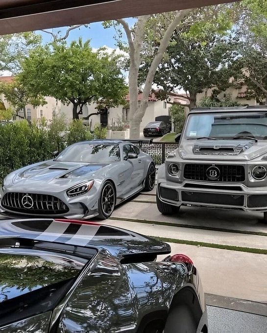 A grey Mercedes-AMG sports car parked next to a grey Mercedes-Benz Brabus G-Wagon in a residential driveway, with the roof of a black car visible in the foreground.