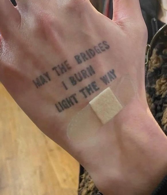 A person's hand is shown displaying a small tattoo reading "MAY THE BRIDGES I BURN LIGHT THE WAY," with a square adhesive bandage placed just below the fresh ink.