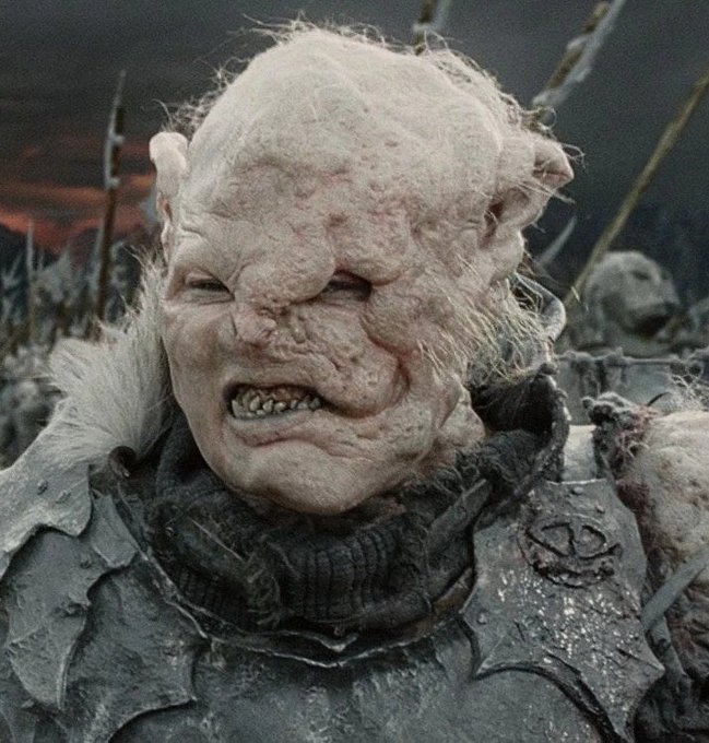 A close-up of an orc-like creature with pale, scarred skin, pointed ears, and a snarling expression, wearing dark armor with fur trim, appearing to be from a fantasy film.