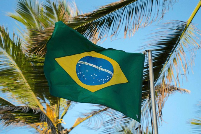 The Brazilian flag waves against a backdrop of palm trees and blue sky, symbolizing the country's new regulatory stance on cryptocurrency exchanges, which mandates a delay of up to 24 hours for large overseas crypto transfers starting in 2027.