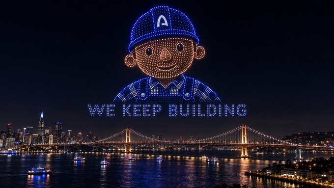 A nighttime drone light show above a city skyline and bridge forms an image of a smiling cartoon worker wearing a hard hat with the letter 'A', accompanied by the text 'WE KEEP BUILDING'.