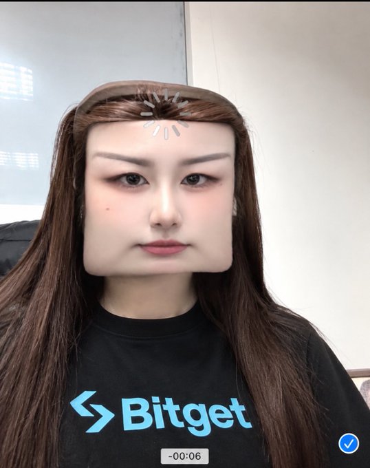 A close-up screenshot of a person wearing a black 'Bitget' t-shirt, with a face filter distorting their features into a square shape.