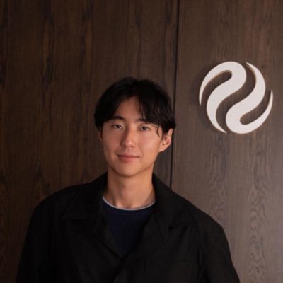 A young man in a black jacket standing in front of a wooden wall with a glowing Sui cryptocurrency logo.