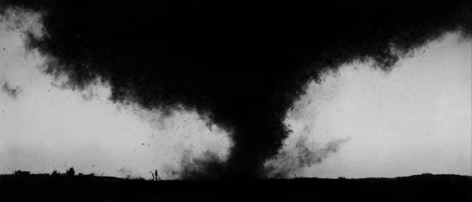 A grainy black and white photograph captures a massive, dark tornado funnel cloud stretching from the sky down to a flat landscape horizon.
