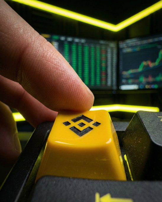 A finger presses a yellow Binance-branded keycap on a mechanical keyboard, with stock or crypto trading charts displayed on dual monitors in the background, visually reinforcing the post's call to action to start trading with Binance.