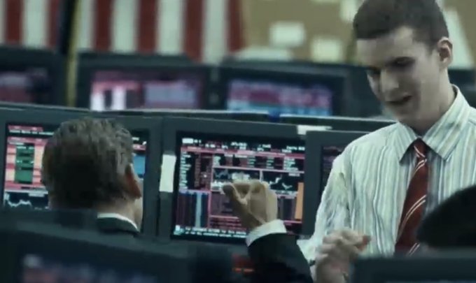 The image shows a man wearing a striped shirt, tie, and glasses standing next to another person who appears to be a computer screen displaying stock market data. The two men are engaged in conversation about the stock market.