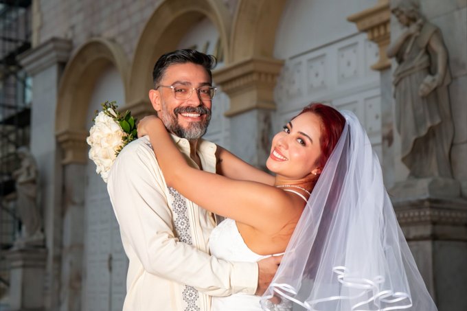 A wedding portrait of a smiling couple embracing; the groom wears glasses and holds a white floral bouquet, while the bride wears a white dress and veil, posed in front of an ornate stone building.