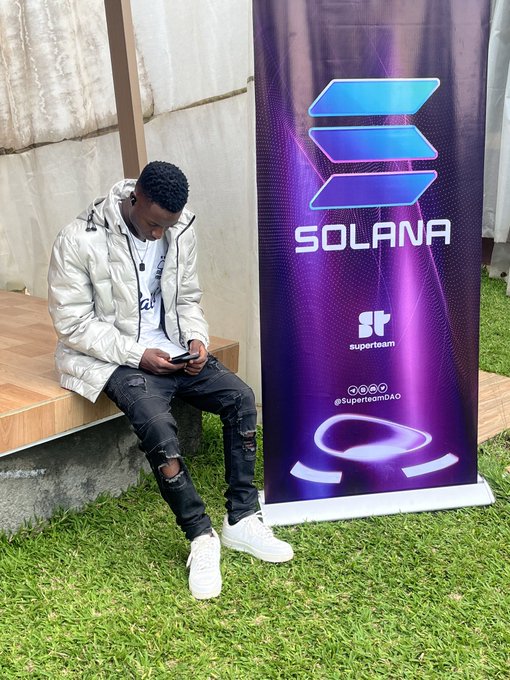 A young man wearing a white puffer jacket and ripped black jeans sits on a wooden bench looking at his phone, next to a purple Solana banner branded with 'Superteam DAO' (@SuperteamDAO).