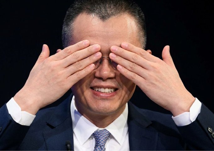 A close-up photo of a smiling man in a dark suit rubbing his eyes with both hands while wearing glasses.