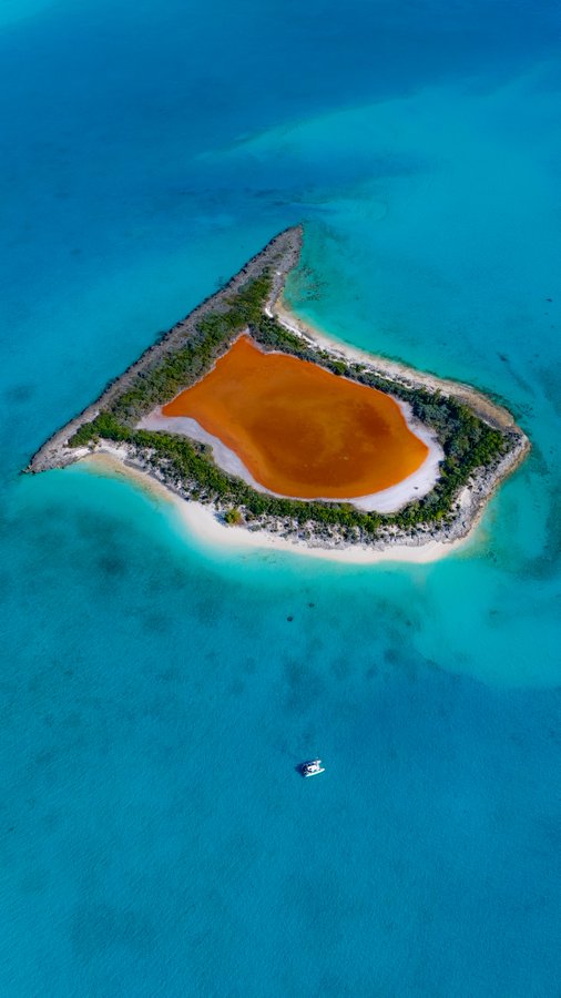 An aerial view of a triangular tropical island with a rust-colored inland lake surrounded by white sandy beaches and turquoise waters.