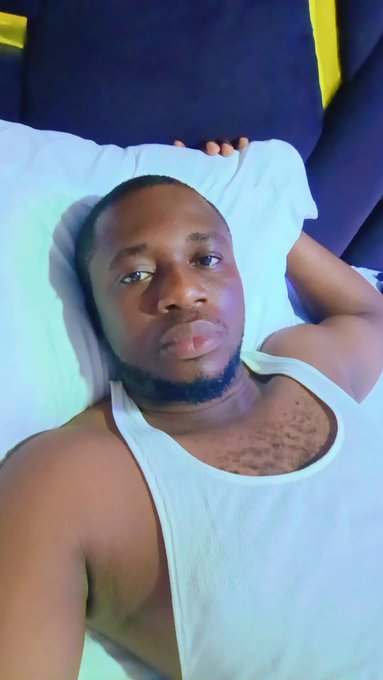 A selfie video screenshot of a young man wearing a white tank top lying in bed with a white pillow, with his arm resting behind his head and a dark blue and yellow background behind him.