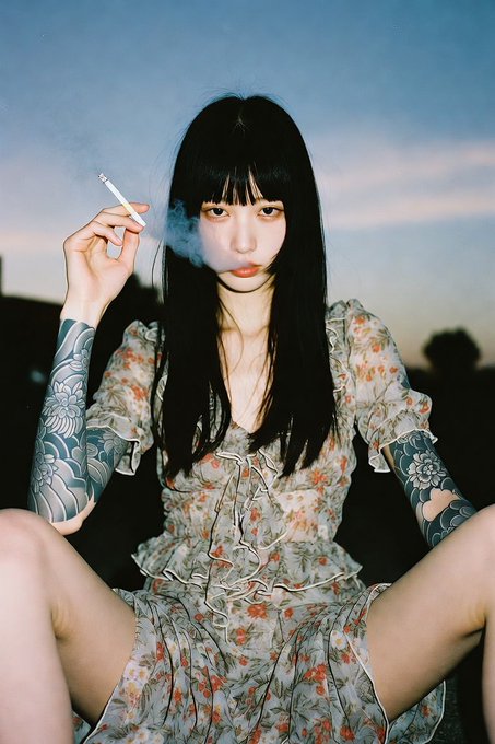 A portrait of a young woman with long black hair and bangs, wearing a floral dress and sporting arm tattoos, sitting outdoors at dusk while smoking a cigarette.