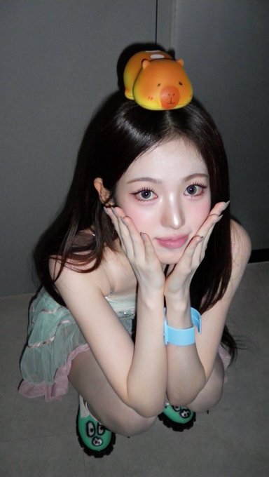 A young woman with large eyes crouching and framing her face with her hands, wearing a small hamster hat and green fuzzy slippers.