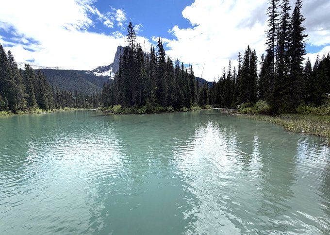 A serene turquoise lake surrounded by evergreen trees with a rocky mountain peak visible in the background under a partly cloudy sky.