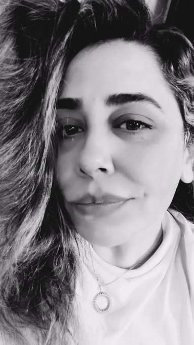 A black-and-white close-up selfie shows a young woman with long, wavy dark hair, wearing a white t-shirt and a delicate circular pendant necklace, gazing softly at the camera. The intimate, grayscale aesthetic reflects the warm personal tone of the post, which celebrates a sale of an artwork with heartfelt appreciation for the buyer.
