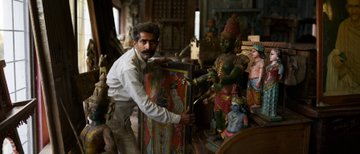A man in a vest stands in a dimly lit room filled with religious statues and artwork, holding what appears to be a colorful icon.