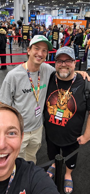Three men pose for a selfie at what appears to be a convention, with one wearing an 'ALF' t-shirt and another in a 'VeeFriends' shirt, with a 'KIDS' Book nook' sign visible in the background.