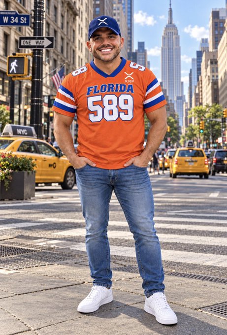 A smiling man wearing an orange 'Florida 589' jersey, blue cap, and jeans stands on a New York City street with yellow taxis and the Empire State Building in the background.