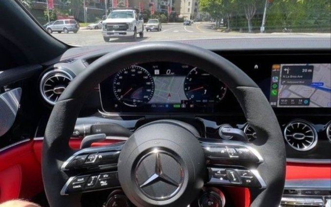 A view from inside a Mercedes-Benz showing the steering wheel, digital dashboard, and navigation screen, with a white truck visible through the windshield.