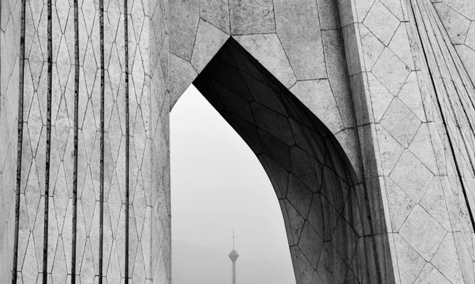 A black and white photograph shows the intricate geometric stone patterns of the Azadi Tower in Tehran, with a tower visible through the archway.